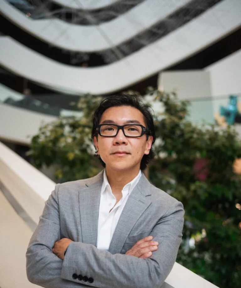 Professional man in a gray blazer with glasses, arms crossed, standing near a railing indoors in a modern atriumcool setting.