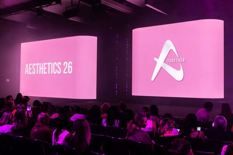 Audience seated in a dark room watching a pink-lit conference stage with two large screens; left reads 'AESTHETICS 26', right shows a white logo and 'TOGETHER'.